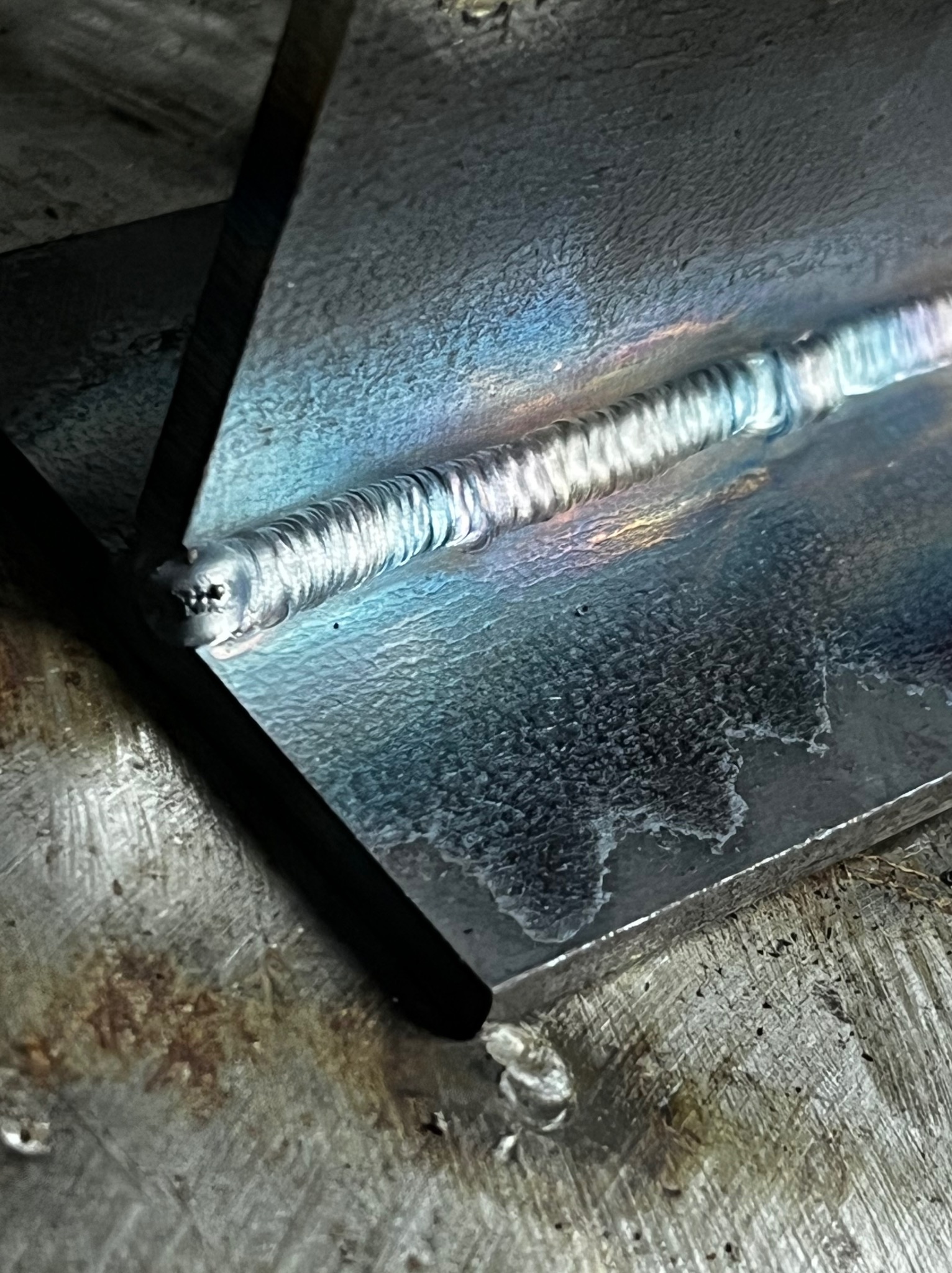 Zbliżenie na świeży spaw dwóch metalowych elementów, widoczne tęczowe przebarwienia na spoinie i powierzchni metalu.