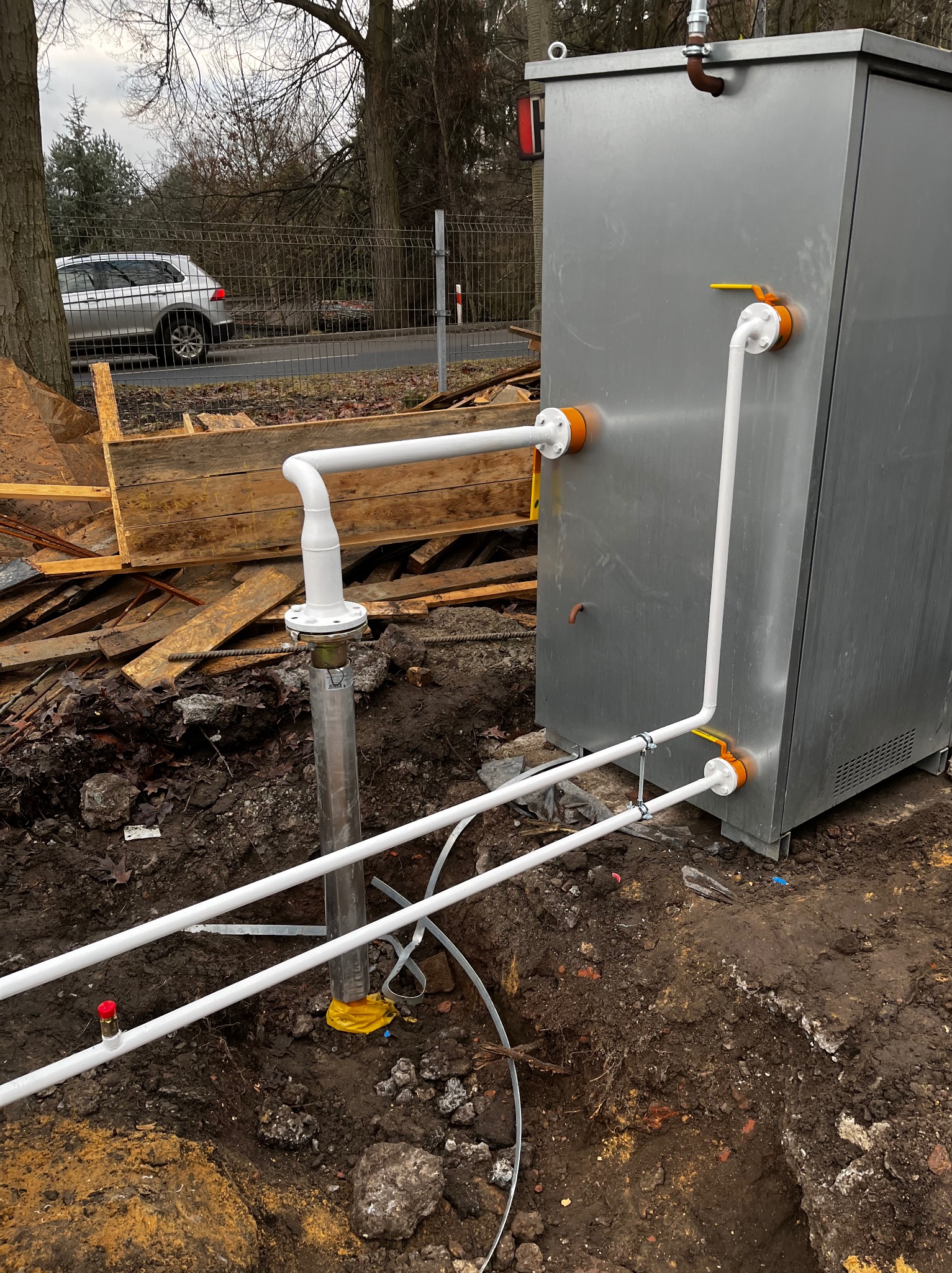 Instalacja gazowa zewnętrzna: metalowa szafa z białymi rurami i żółtymi zaworami, częściowo wkopana w ziemię, widoczne wykopaliska i elementy budowlane w tle.