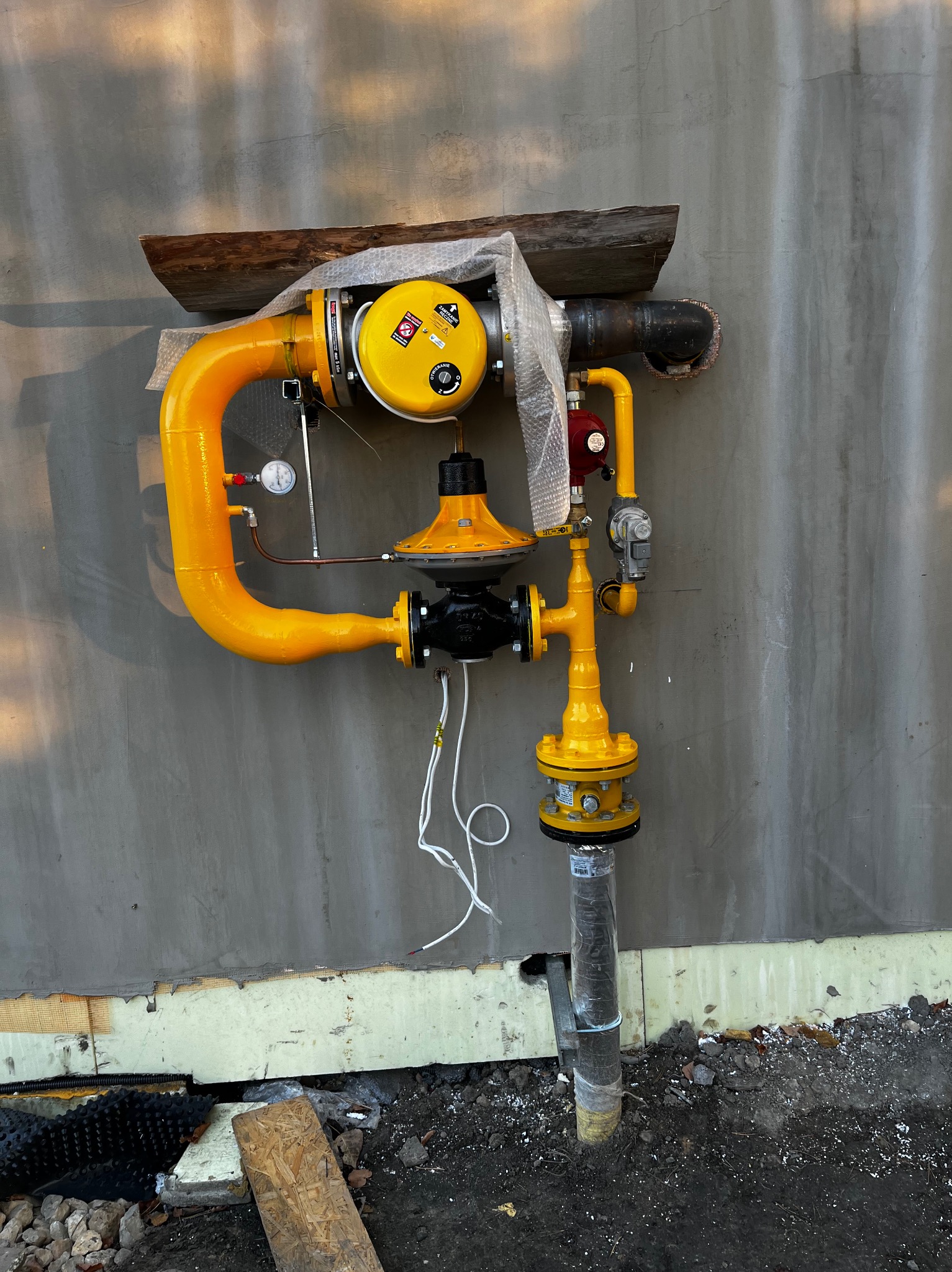 Kompletna, żółta instalacja gazowa zamontowana na szarej ścianie zewnętrznej budynku, z widocznymi elementami zabezpieczającymi i manometrem, na tle częściowo wykończonej elewacji.