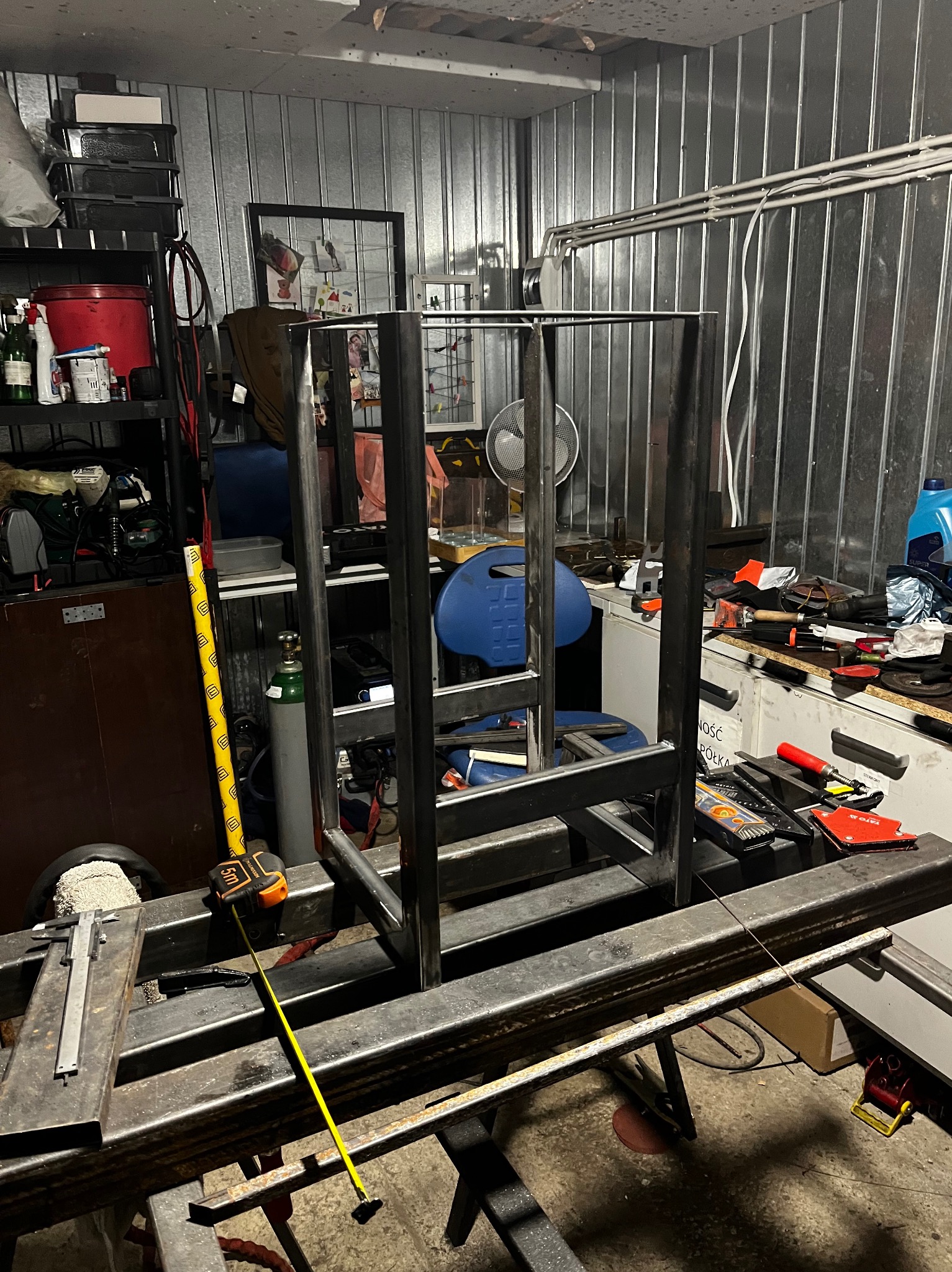 Konstrukcja metalowa w trakcie spawania w warsztacie, widoczny spawacz, butla z gazem, miara i narzędzia na stole roboczym.