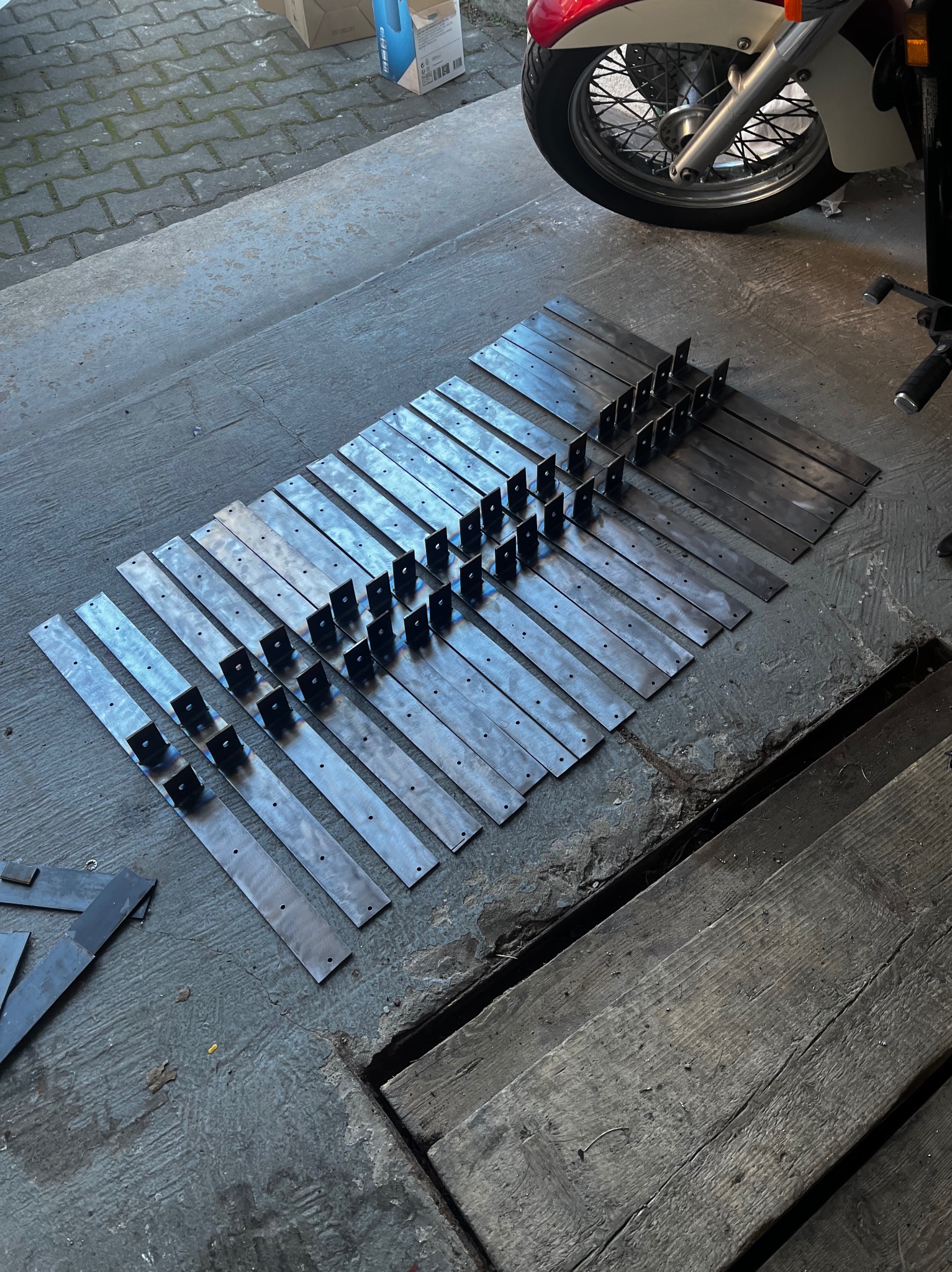 Równy rząd metalowych płaskowników z przyspawanymi uchwytami, ułożonych na betonowej podłodze garażu obok motocykla, gotowych do montażu.