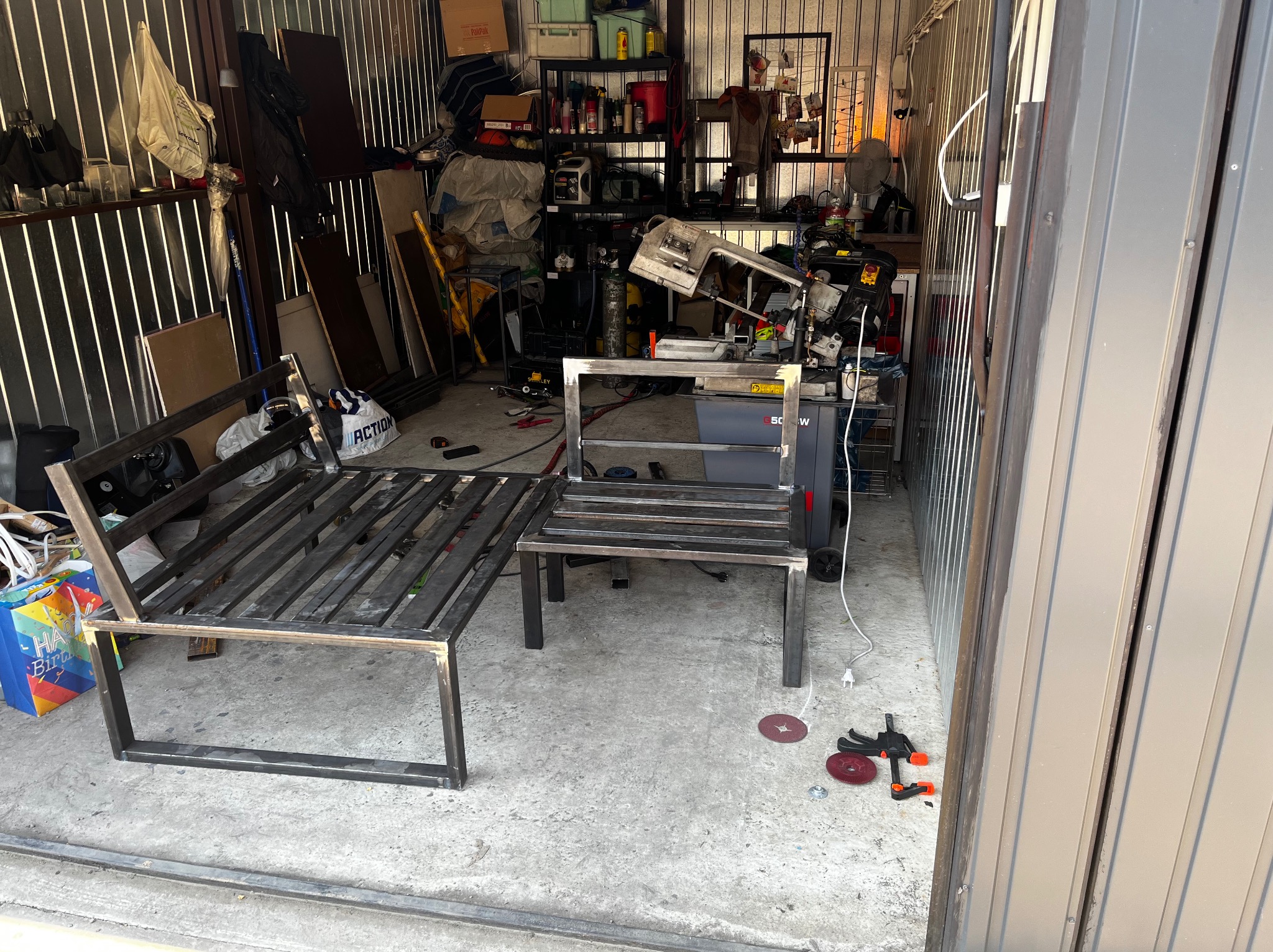 Dwie metalowe konstrukcje w warsztacie, prawdopodobnie ramy meblowe, w surowym stanie, z widocznymi śladami spawania, w tle narzędzia i materiały warsztatowe.