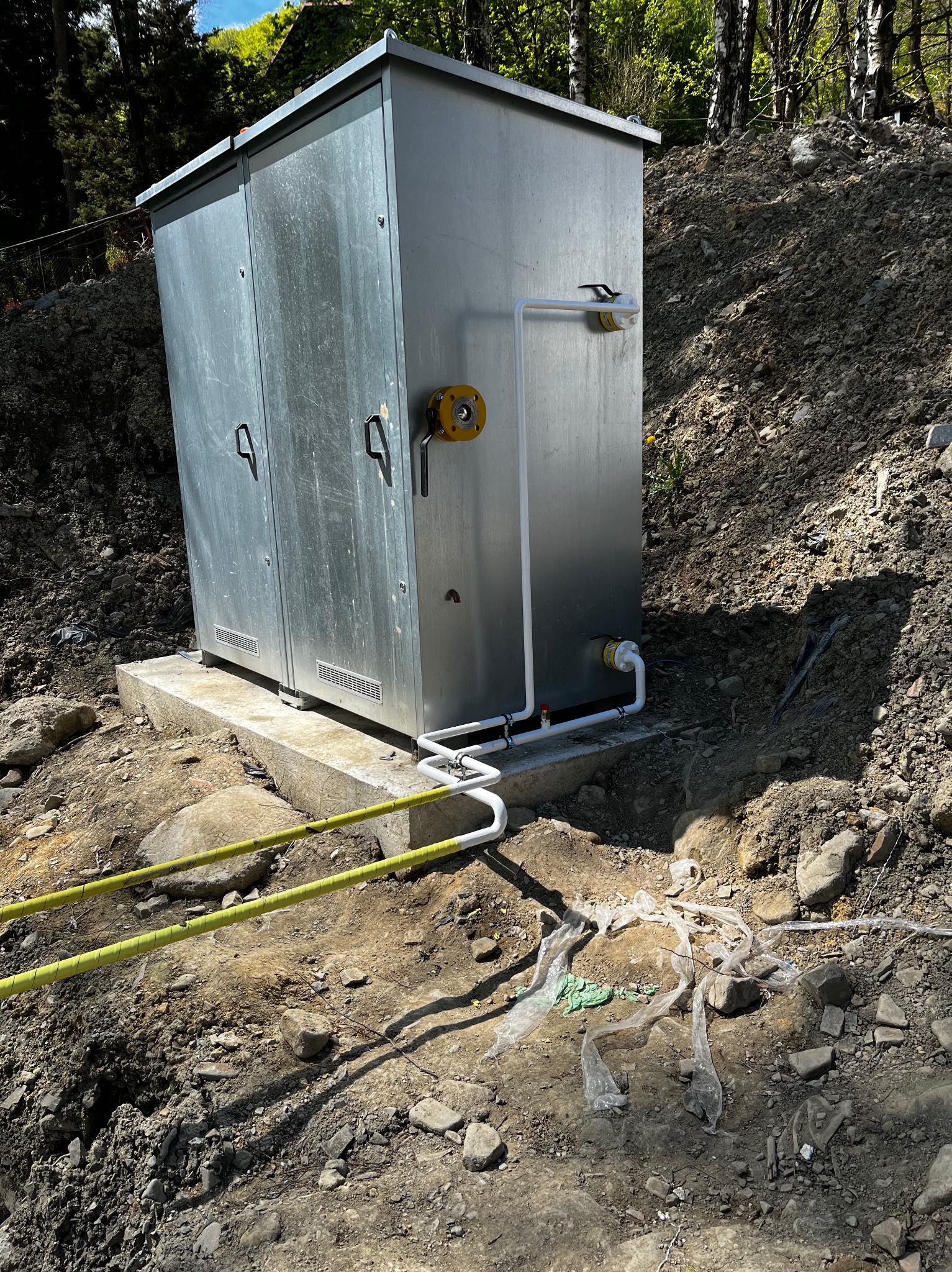 Metalowa szafa instalacyjna z rurami na betonowej podstawie, umieszczona na nierównym terenie ze świeżo usypaną ziemią i żółtymi barierkami ostrzegawczymi.