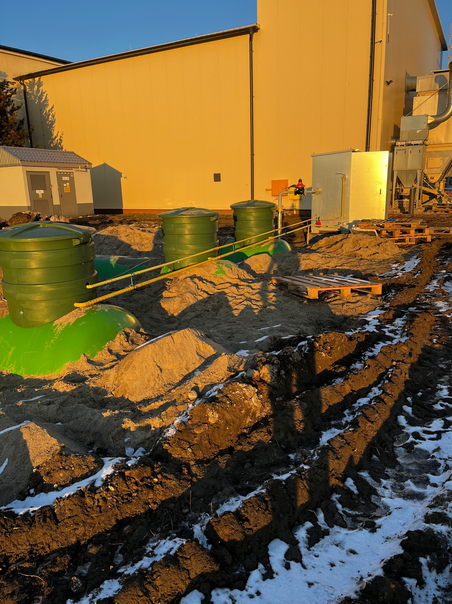 Instalacja trzech zielonych zbiorników zakopanych w piasku, połączonych żółtymi rurami, z budynkiem przemysłowym w tle, na częściowo ośnieżonym terenie.