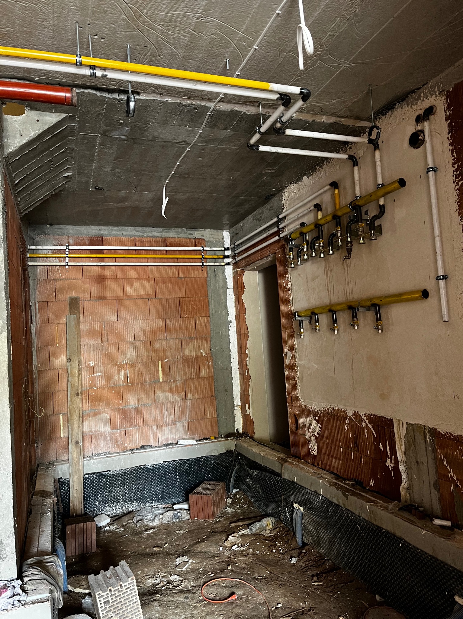 Wnętrze w trakcie remontu z widocznymi instalacjami gazowymi i wodnymi, ściany częściowo w surowym stanie, widoczne rury w kolorach żółtym, białym i czerwonym, zamocowane do ściany i sufitu...