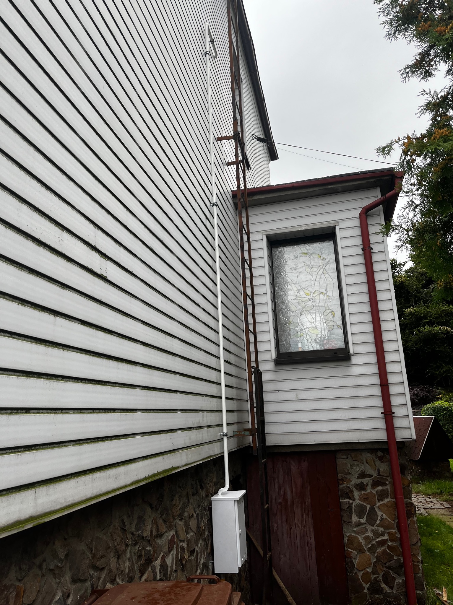 Biała rura gazowa pionowo przymocowana do ściany domu z sidingiem, obok drabina metalowa i okno z witrażem.