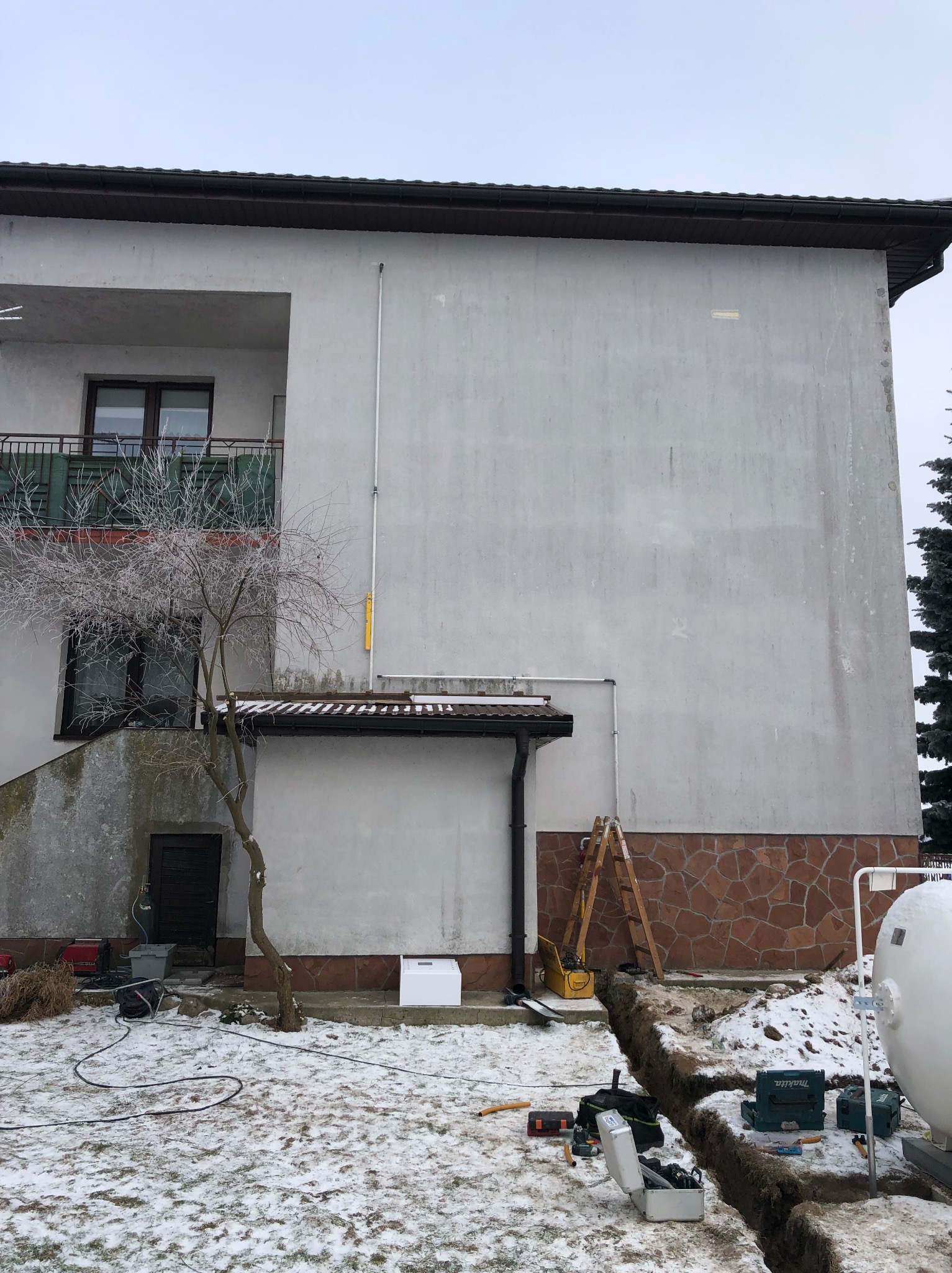 Instalacja gazowa zewnętrzna w Sosnowcu: Wykop podziemny, biały zbiornik LPG, rury gazowe prowadzone po elewacji budynku, drabina oparta o ścianę, skrzynki narzędziowe i sprzęt roboczy widoczne...