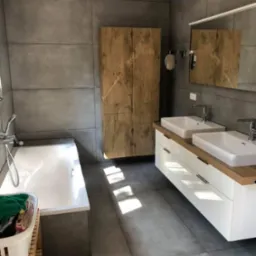 Nowoczesna łazienka z szarymi płytkami wielkoformatowymi, drewnianą szafką, podwójną umywalką na białej szafce z drewnianym blatem i wanną z praniem w koszu.