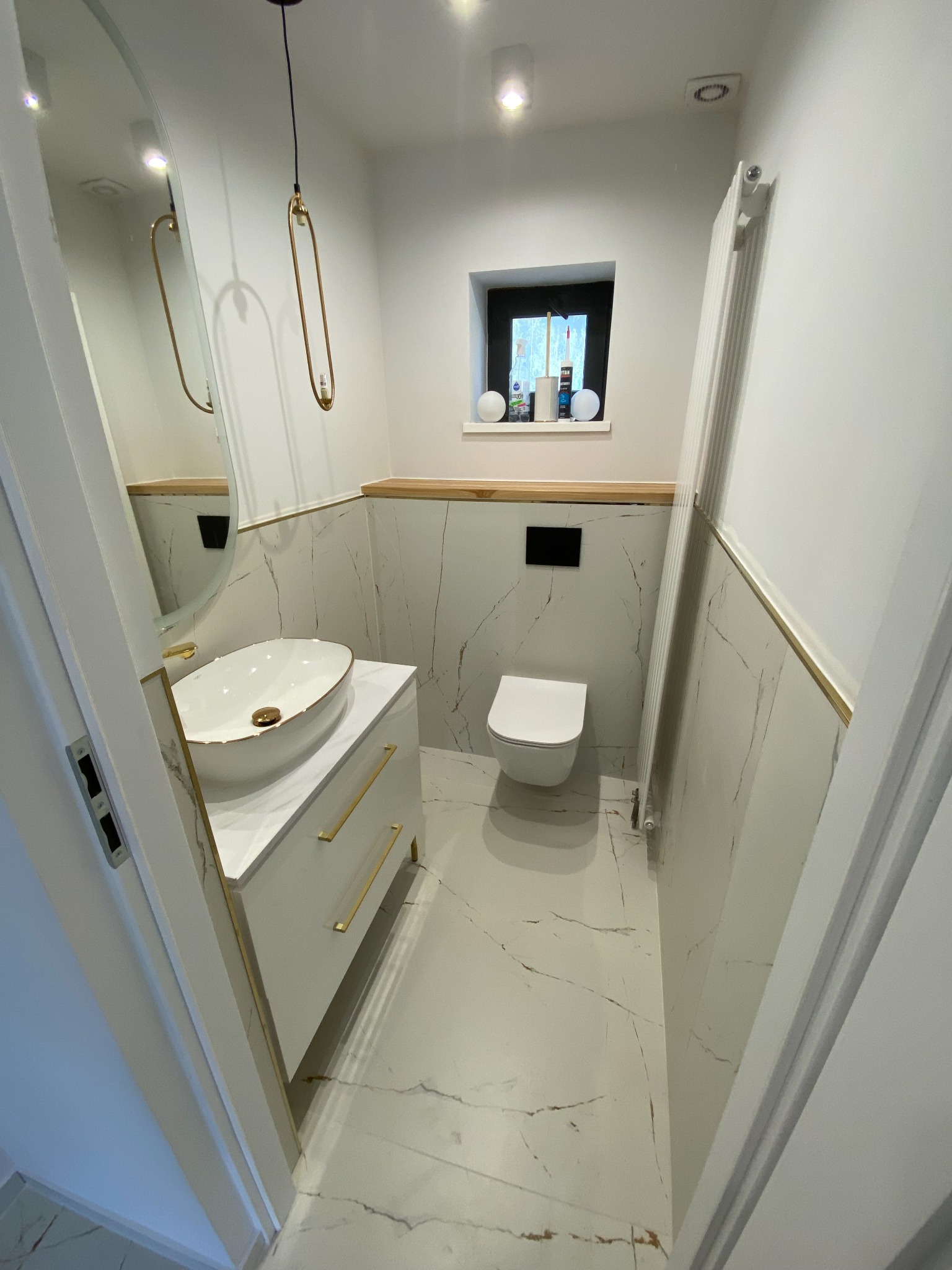 Nowoczesna łazienka w bieli i marmurze z owalnym lustrem, umywalką nablatową w kolorze ecru ze złotymi akcentami i wiszącą lampą w kształcie podłużnej elipsy