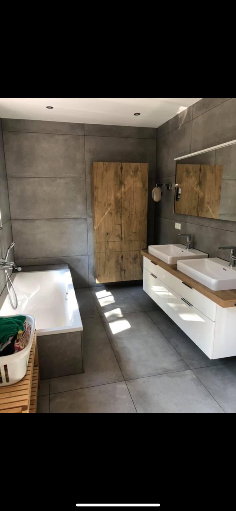 Nowoczesna łazienka z szarymi płytkami wielkoformatowymi, drewnianą szafką, podwójną umywalką na białej szafce z drewnianym blatem i wanną z praniem w koszu.