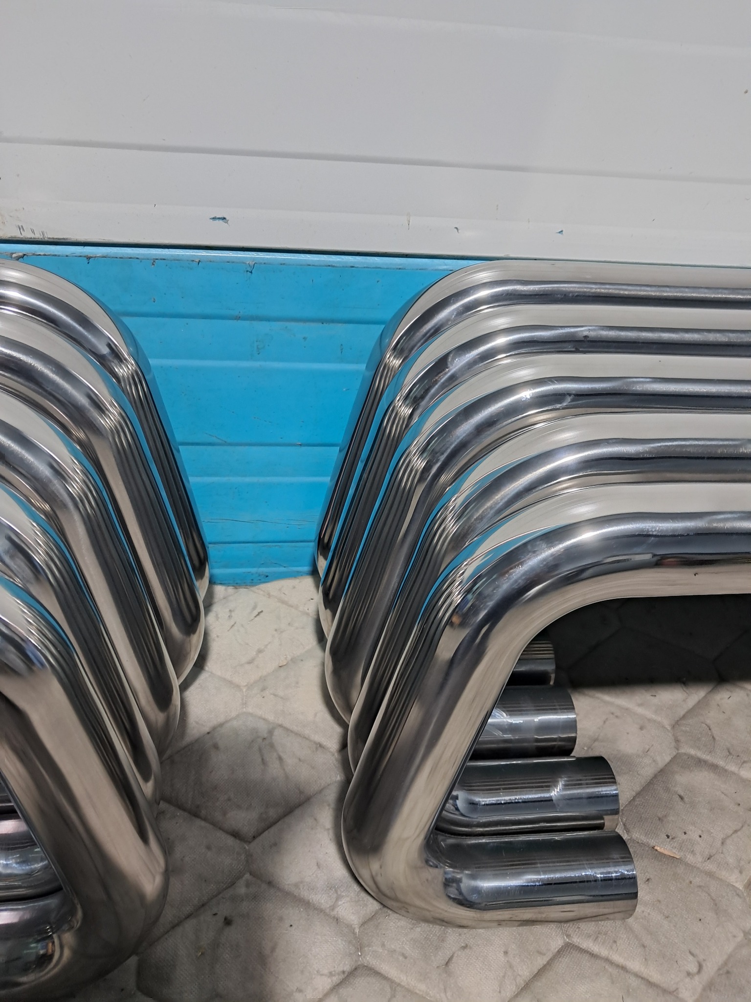 Stos polerowanych, giętych rur ze stali nierdzewnej, ułożonych na szarej, wzorzystej powierzchni, z niebieską i białą ścianą w tle.