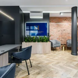 Eleganckie biuro agencji nieruchomości z nowoczesnym biurkiem, welurowymi krzesłami, ścianą z czerwonej cegły i podświetlanym logo firmy nad roślinami w drewnianej donicy.