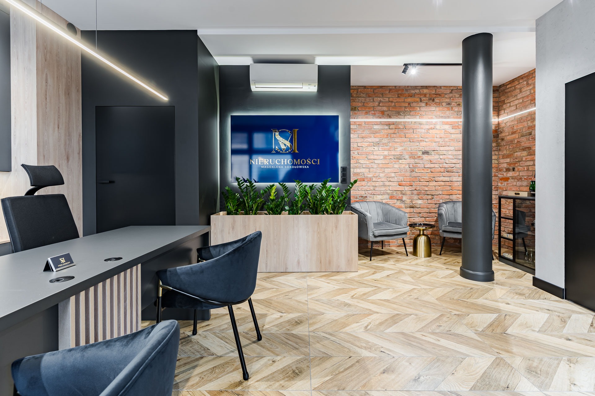 Eleganckie biuro agencji nieruchomości z nowoczesnym biurkiem, welurowymi krzesłami, ścianą z czerwonej cegły i podświetlanym logo firmy nad roślinami w drewnianej donicy.