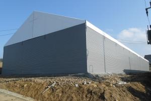 Duża, szara hala z białym dachem, wykonana z blachy trapezowej, widoczna z zewnątrz w słoneczny dzień, z ziemią i skarpą w tle.