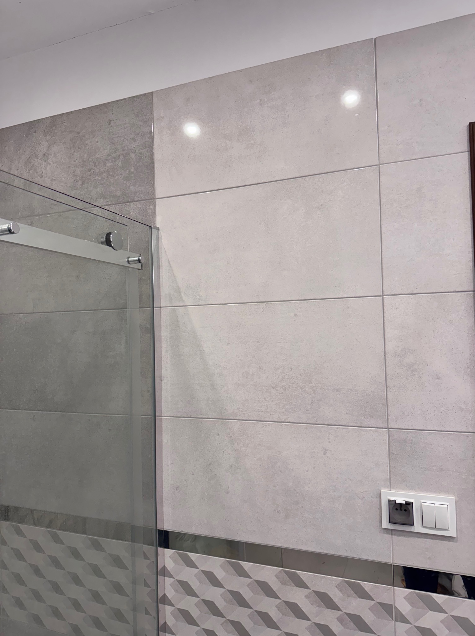 Wykończona łazienka z szarą, geometryczną mozaiką u dołu, jasnymi płytkami na ścianach i szklaną kabiną prysznicową. Widoczny włącznik światła.