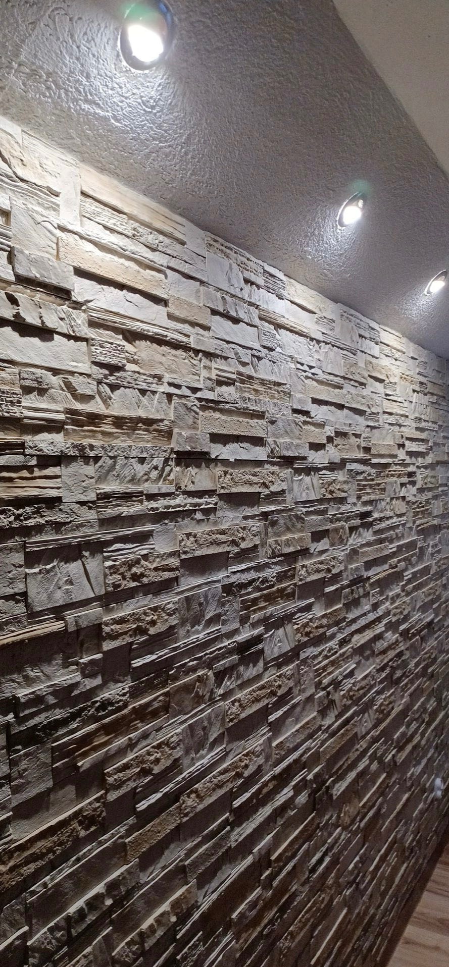 Ściana wykończona dekoracyjnym kamieniem w odcieniach beżu i brązu, oświetlona trzema punktowymi lampami sufitowymi. Teksturowana powierzchnia sufitu.