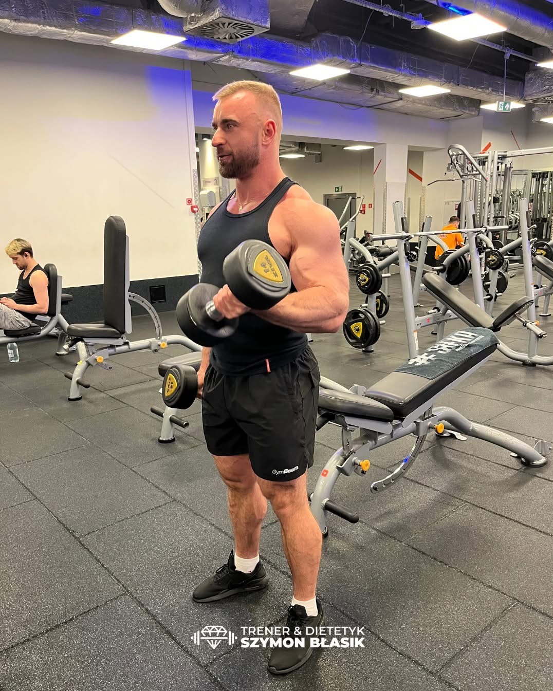 Mężczyzna z krótkimi włosami i brodą, w czarnym stroju sportowym, ćwiczy z hantlą w siłowni, widoczny profil i napięte mięśnie ramion, w tle sprzęt do ćwiczeń i inni ćwiczący.