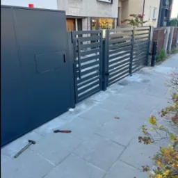 Nowoczesna, antracytowa brama wjazdowa z poziomymi panelami, skrzynka na listy wbudowana w słupek, na chodniku leżą młotek i klucz nastawny.