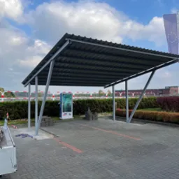 Zadaszenie metalowe nad stanowiskiem ładowania pojazdów elektrycznych, widoczne kostki brukowe przygotowane do montażu, w tle flaga Unii Europejskiej i budynek.