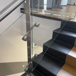 POLER-MET MAREK UDZIK - Szklana balustrada ze stalowymi elementami mocującymi, widok z góry na schody z czarnymi i drewnianymi stopniami, w tle widoczna osoba w odblaskowej kamizelce.