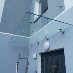 POLER-MET MAREK UDZIK - Szklany daszek nad wejściem do budynku, wsparty na metalowych wspornikach, widoczne drabiny sugerujące prace montażowe.