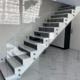 Nowoczesne schody z białymi stopniami i szarą powierzchnią do chodzenia, zabezpieczone szklaną balustradą mocowaną na metalowych uchwytach, na tle marmurowej podłogi i ściany z refleksami światła.