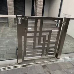 Nowoczesna brama ze stali nierdzewnej z panelem dostępu numerycznego, wkomponowana w szklane ogrodzenie na tle szarej kostki brukowej i jasnej elewacji budynku.