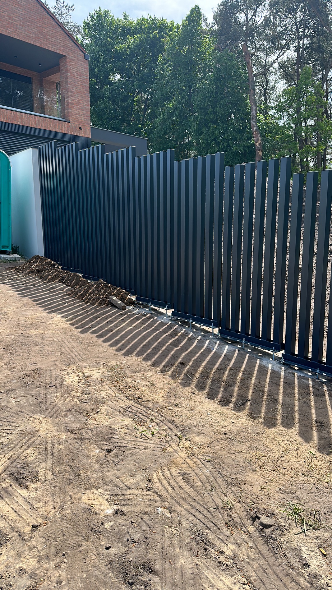Nowoczesne, pionowe ogrodzenie z ciemnych metalowych paneli przy posesji z ceglanym domem, widoczne ślady prac ziemnych i tymczasowa toaleta.