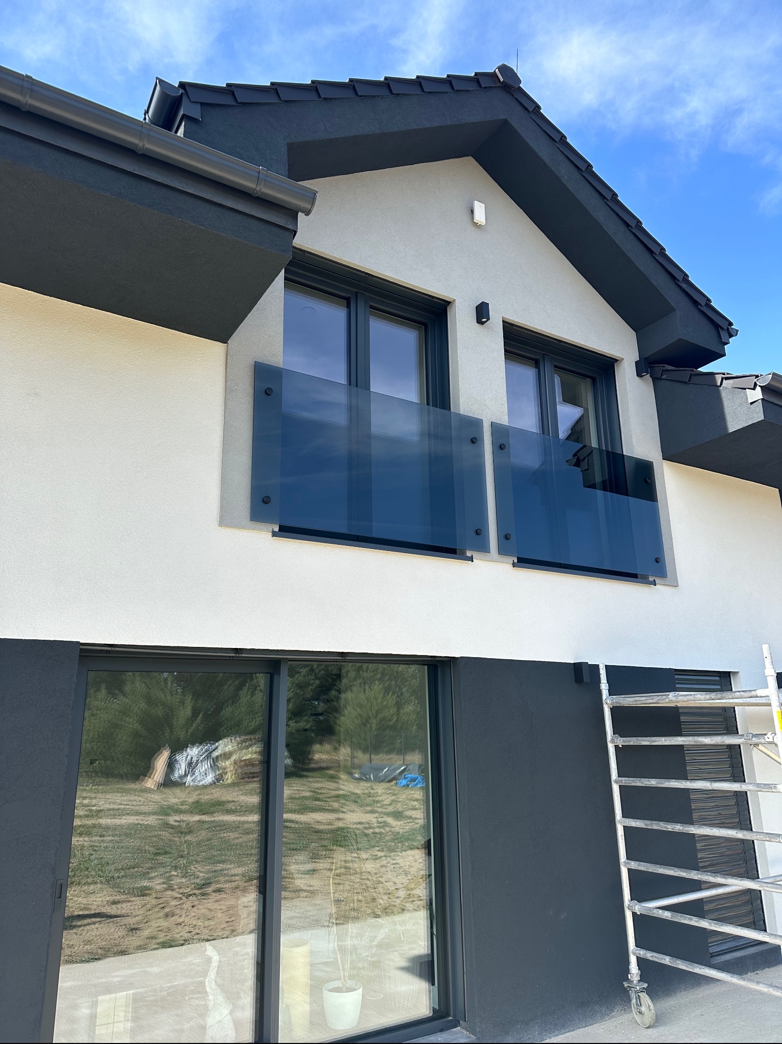 Nowoczesny dom z szarymi detalami, balkon z ciemnymi szklanymi panelami i rusztowanie w tle, sugerujące trwające prace wykończeniowe.