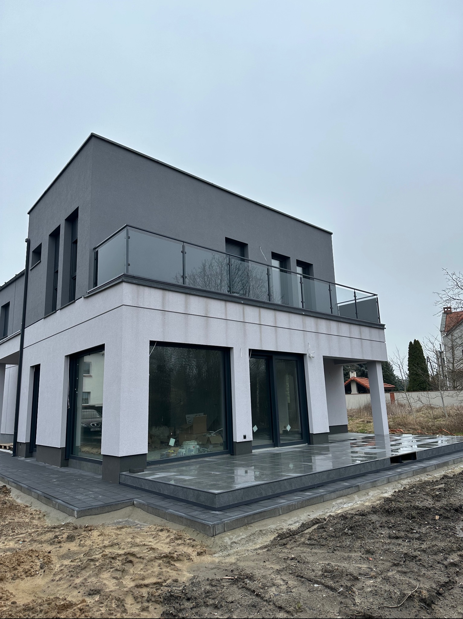 Nowoczesny dom w Czarnkowie z szarą elewacją i szklaną balustradą na balkonie, widok z poziomu gruntu na częściowo wykończony taras z szarych płyt.