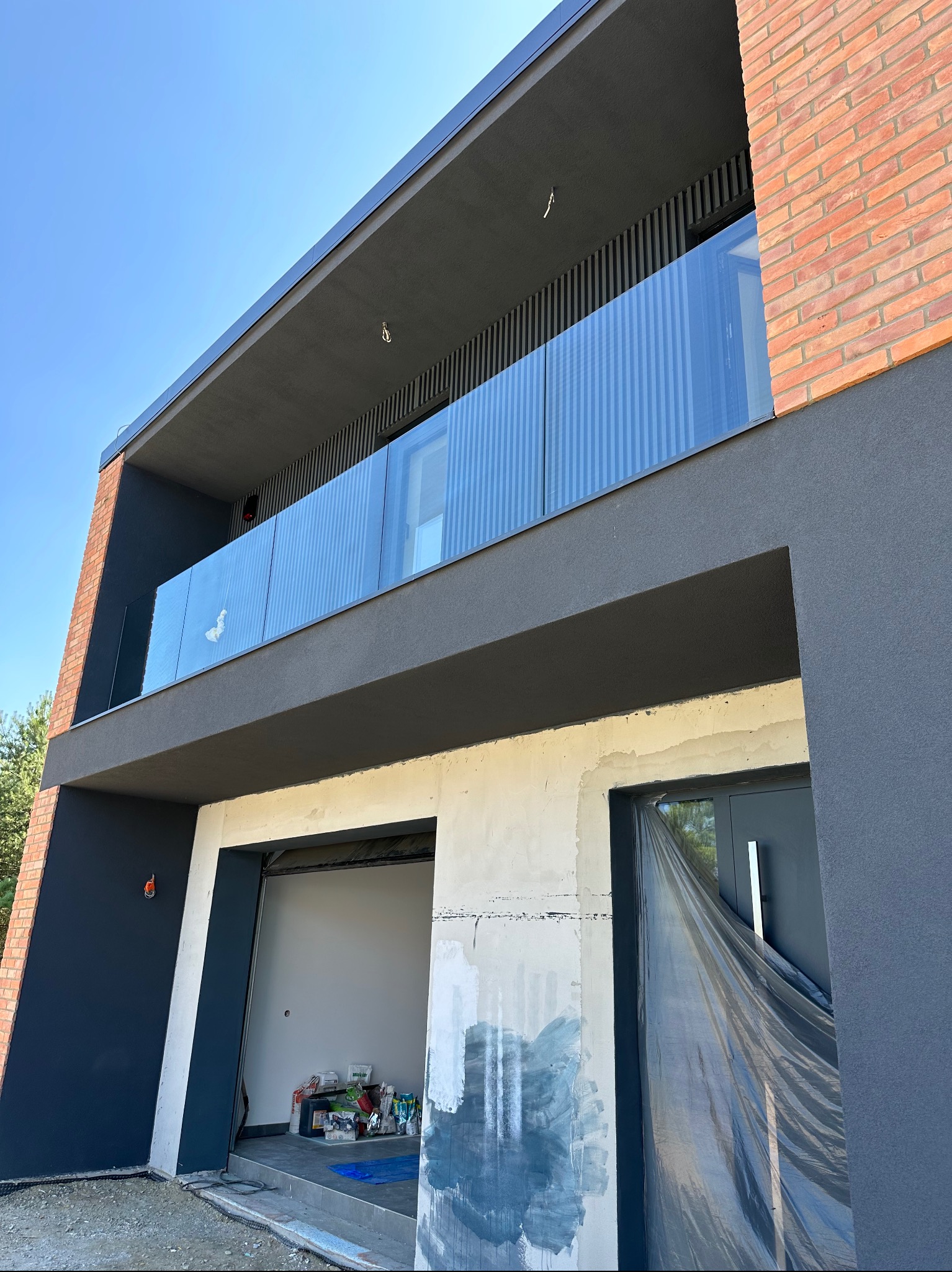 Nowoczesny budynek z balkonem zabezpieczonym szkłem, widok z dołu na elewację w trakcie wykończenia, z widocznymi materiałami budowlanymi.