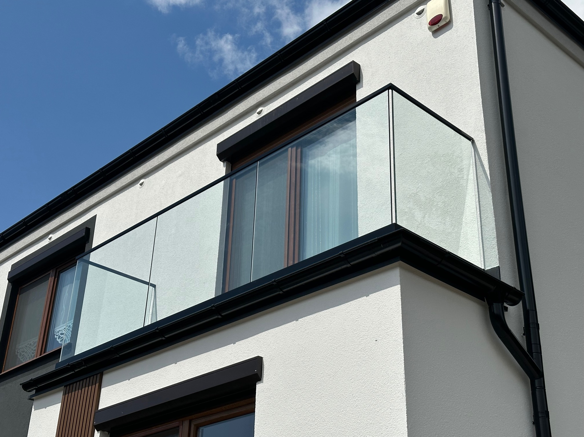 Nowoczesny balkon z przezroczystą, szklaną balustradą, czarne ramy okienne i orynnowanie na tle białej, teksturowanej elewacji budynku.