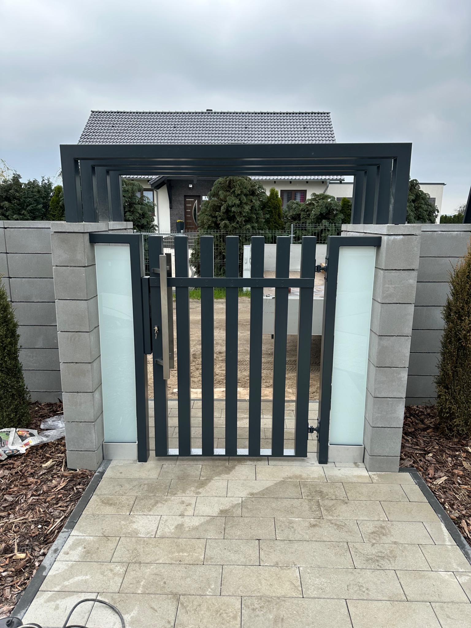 Nowoczesna, antracytowa furtka z pionowymi szczeblami i mlecznym szkłem, osadzona w betonowych słupkach z pergolą, prowadząca do domu z ogrodem.