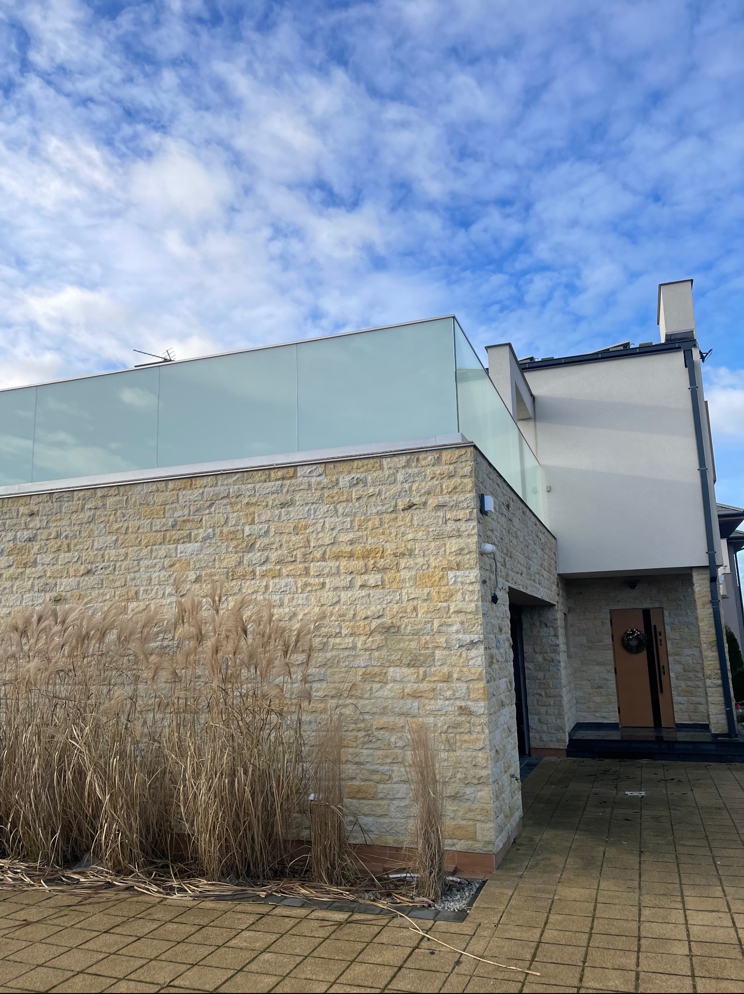 Nowoczesny dom z kamienną elewacją i szklaną balustradą na tarasie, widok z zewnątrz w słoneczny dzień.