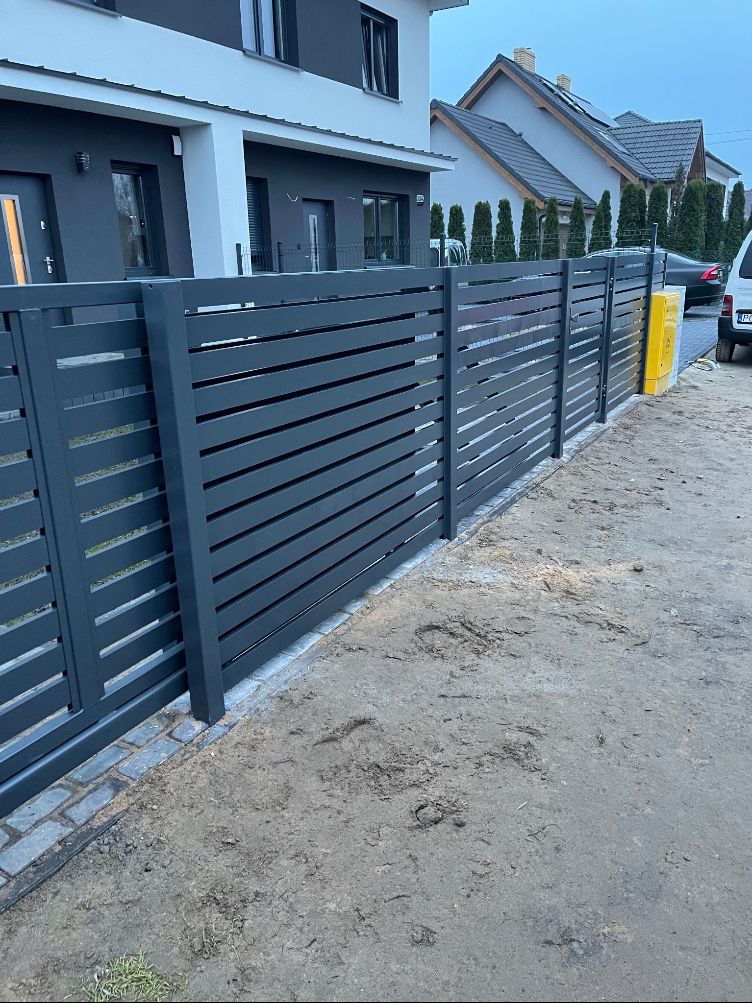 Nowoczesne ogrodzenie panelowe w kolorze grafitowym, z poziomymi listwami, oddzielające posesję od drogi gruntowej, z widocznym fragmentem domu i samochodu w tle.