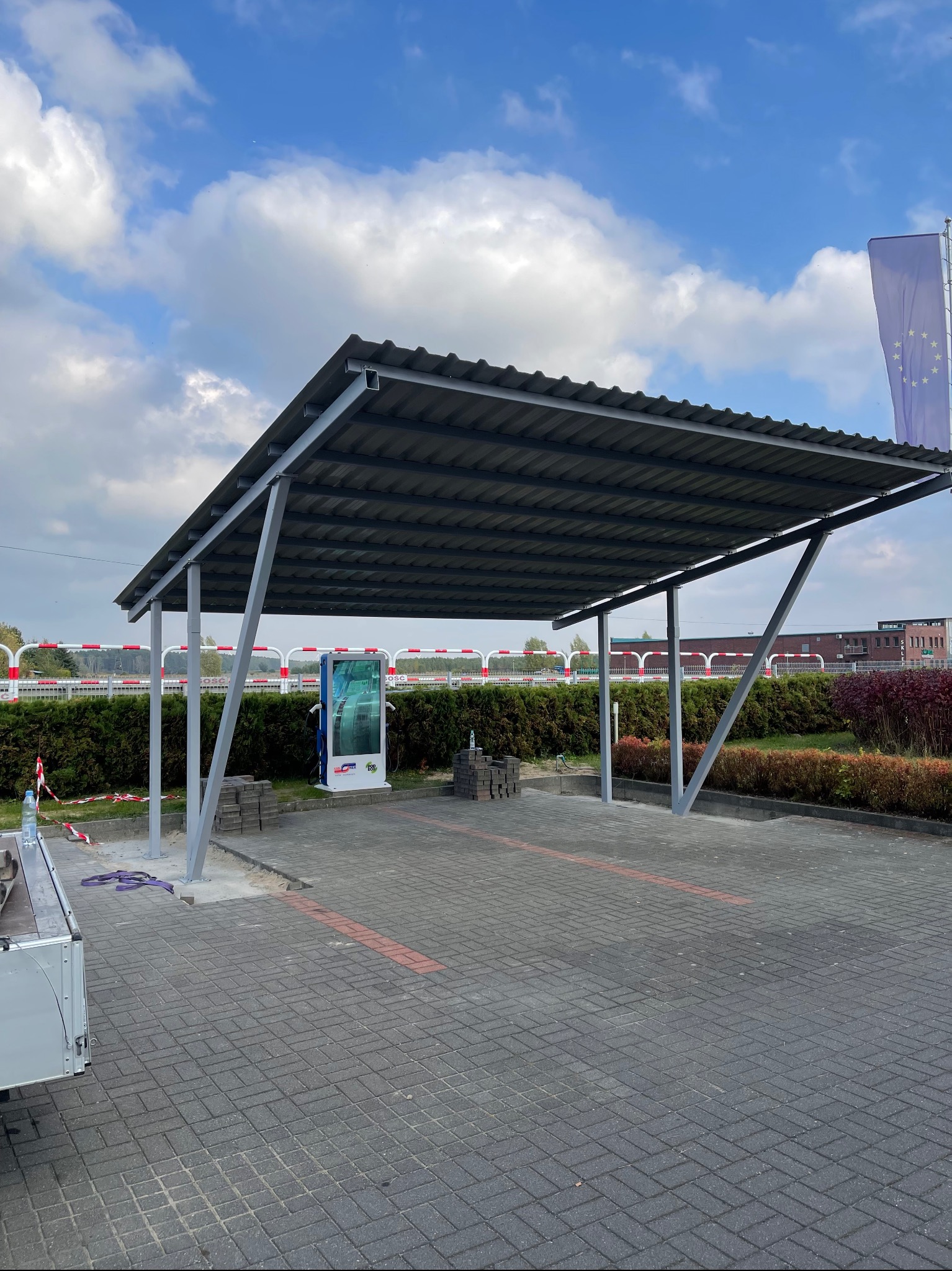 Zadaszenie metalowe nad stanowiskiem ładowania pojazdów elektrycznych, widoczne kostki brukowe przygotowane do montażu, w tle flaga Unii Europejskiej i budynek.
