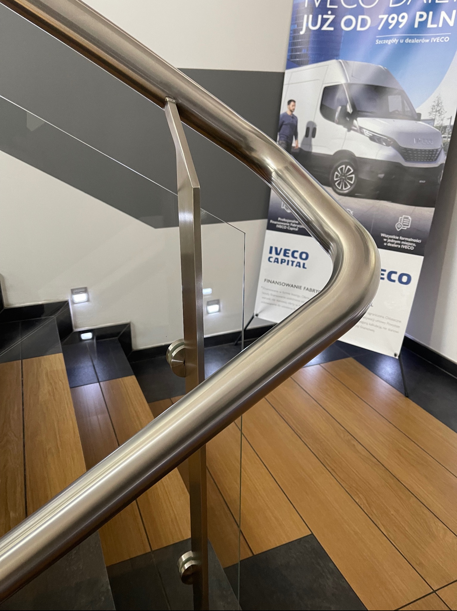 Szklana balustrada z nierdzewną poręczą na schodach wykończonych drewnem i czarnym gresem, w tle reklama Iveco.