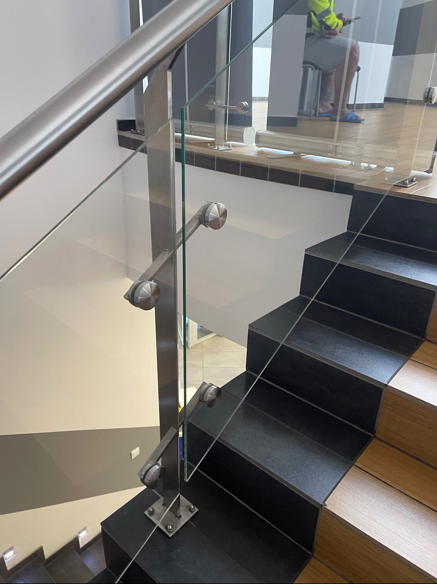 Szklana balustrada ze stalowymi elementami mocującymi, widok z góry na schody z czarnymi i drewnianymi stopniami, w tle widoczna osoba w odblaskowej kamizelce.