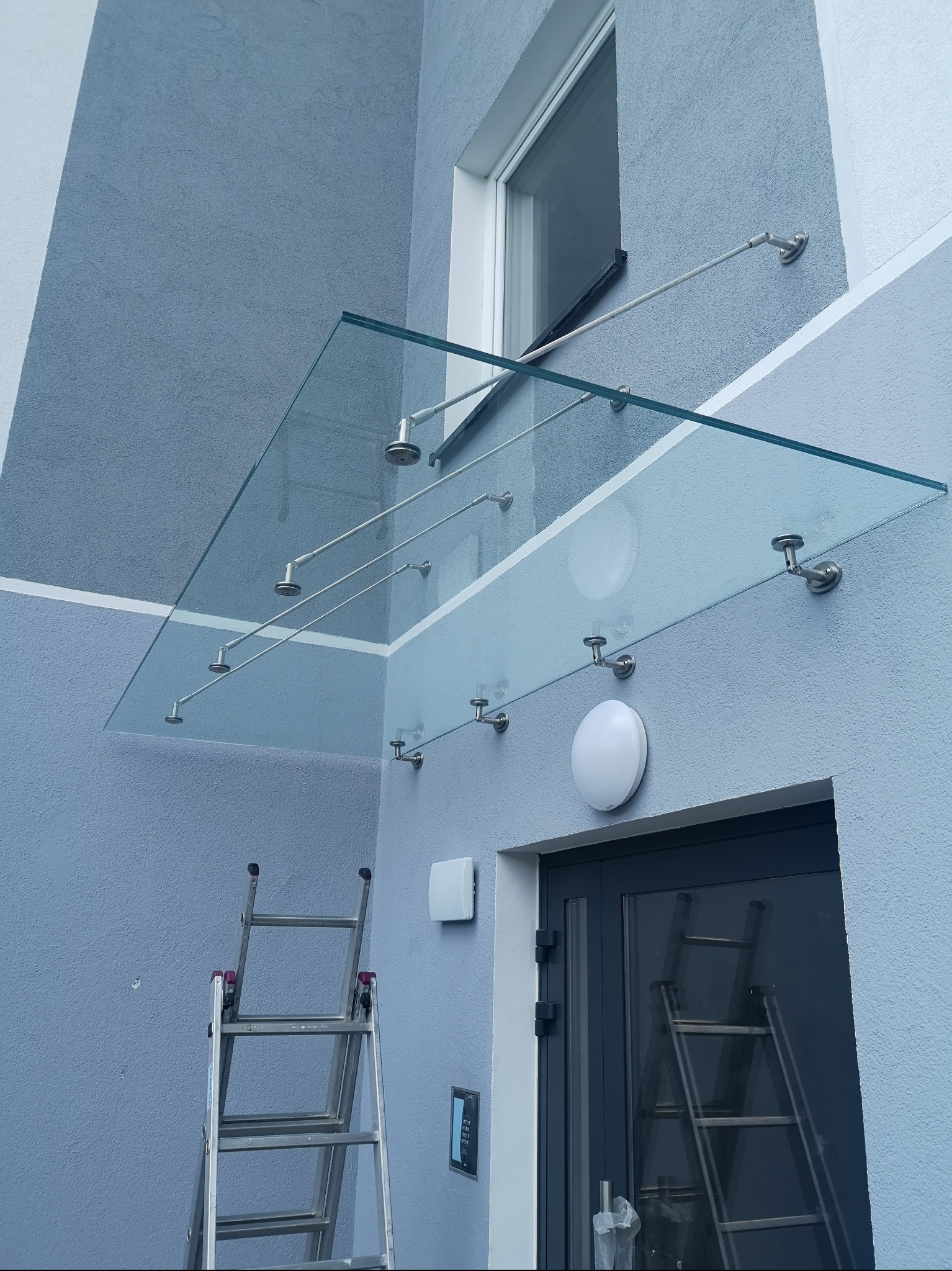 Szklany daszek nad wejściem do budynku, wsparty na metalowych wspornikach, widoczne drabiny sugerujące prace montażowe.