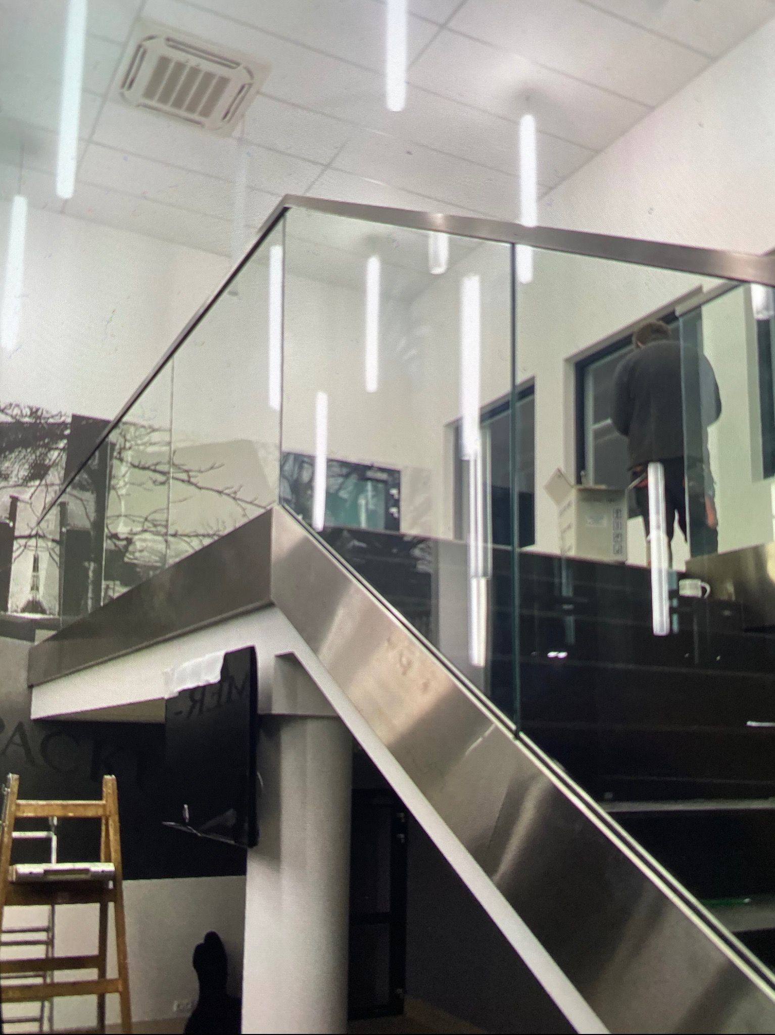 Wnętrze z zamontowaną szklaną balustradą na schodach, widoczny pracownik w trakcie montażu, drabina oparta o ścianę, kartony i narzędzia w tle.