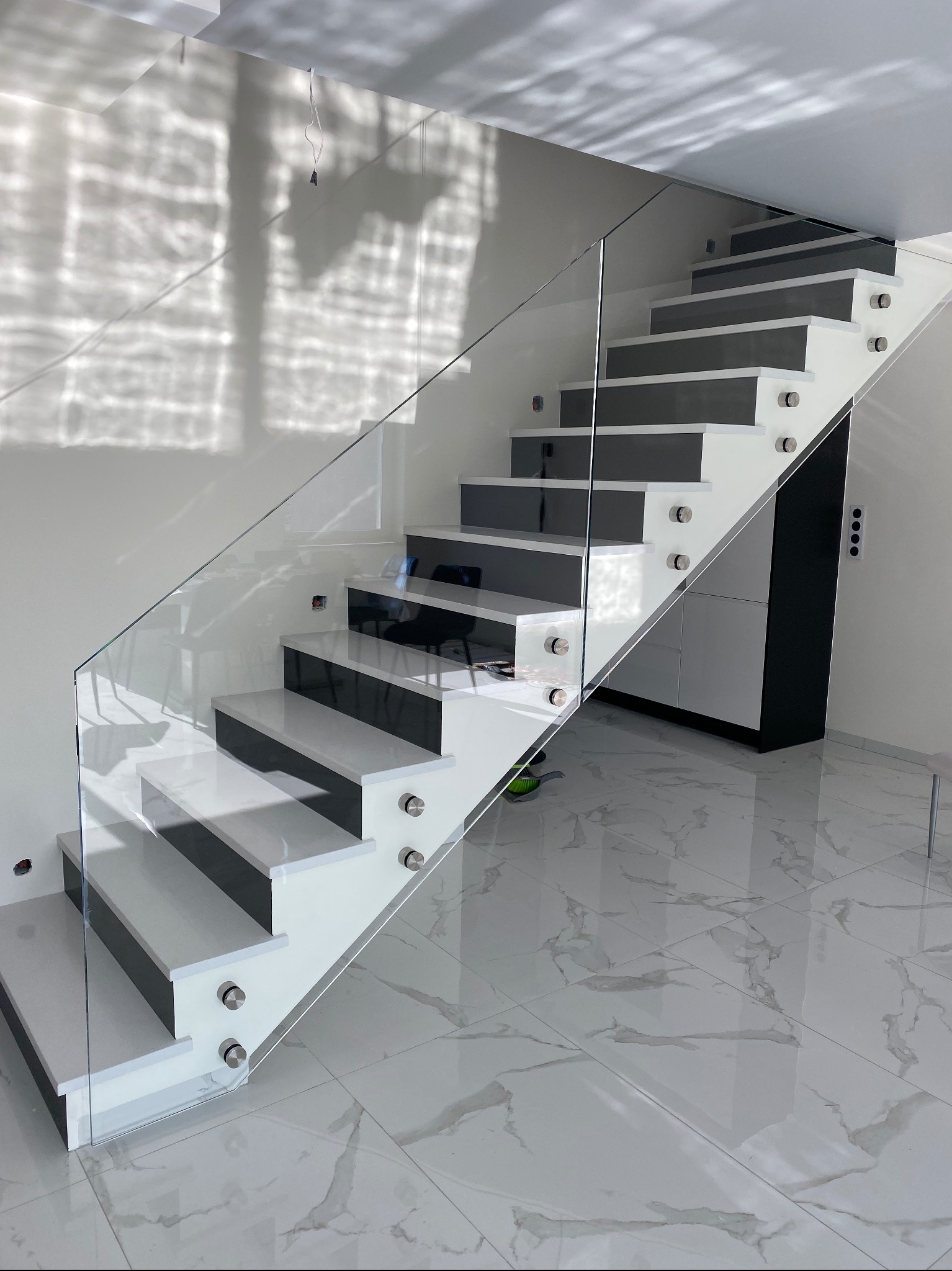 Nowoczesne schody z białymi stopniami i szarą powierzchnią do chodzenia, zabezpieczone szklaną balustradą mocowaną na metalowych uchwytach, na tle marmurowej podłogi i ściany z refleksami światła.