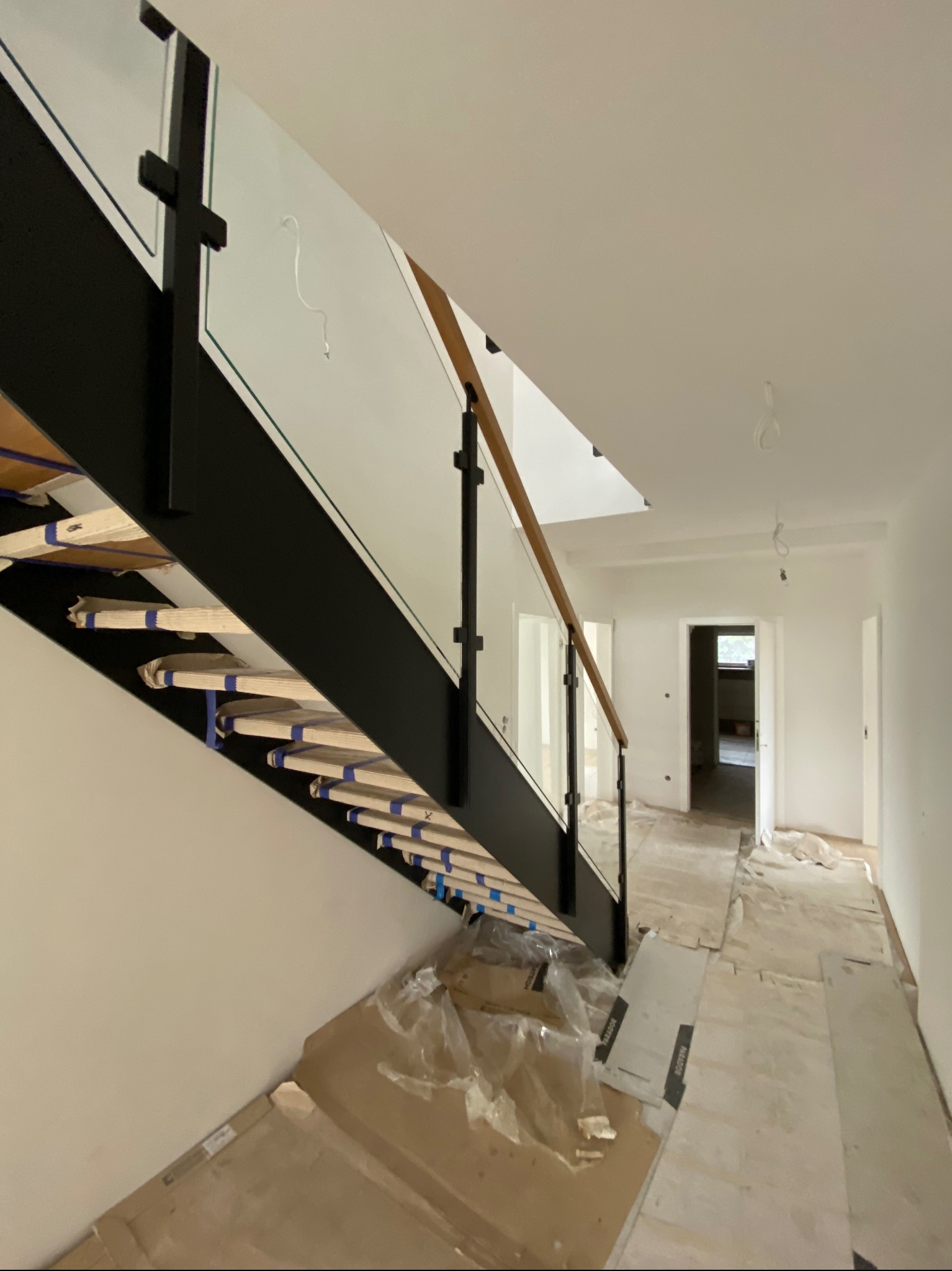 Wnętrze w trakcie wykańczania: schody z czarną metalową konstrukcją, drewnianymi stopniami zabezpieczonymi folią i szkłem, widok na korytarz z otwartymi drzwiami do innych pomieszczeń.