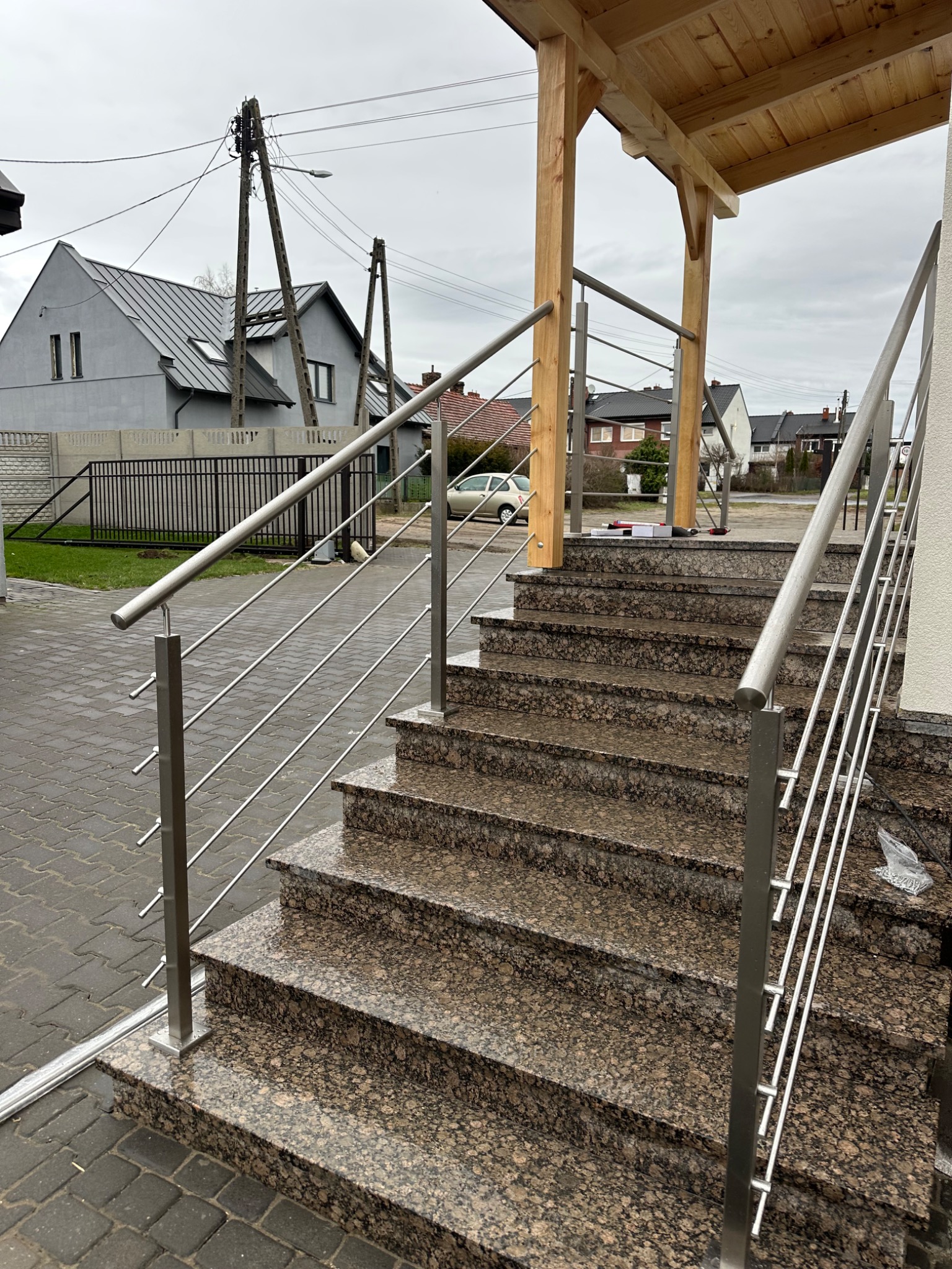 Zewnętrzne schody z brązowego, nakrapianego granitu z nowoczesną, metalową balustradą ze stali nierdzewnej z wypełnieniem linkami, prowadzące do drewnianego zadaszenia ganku.