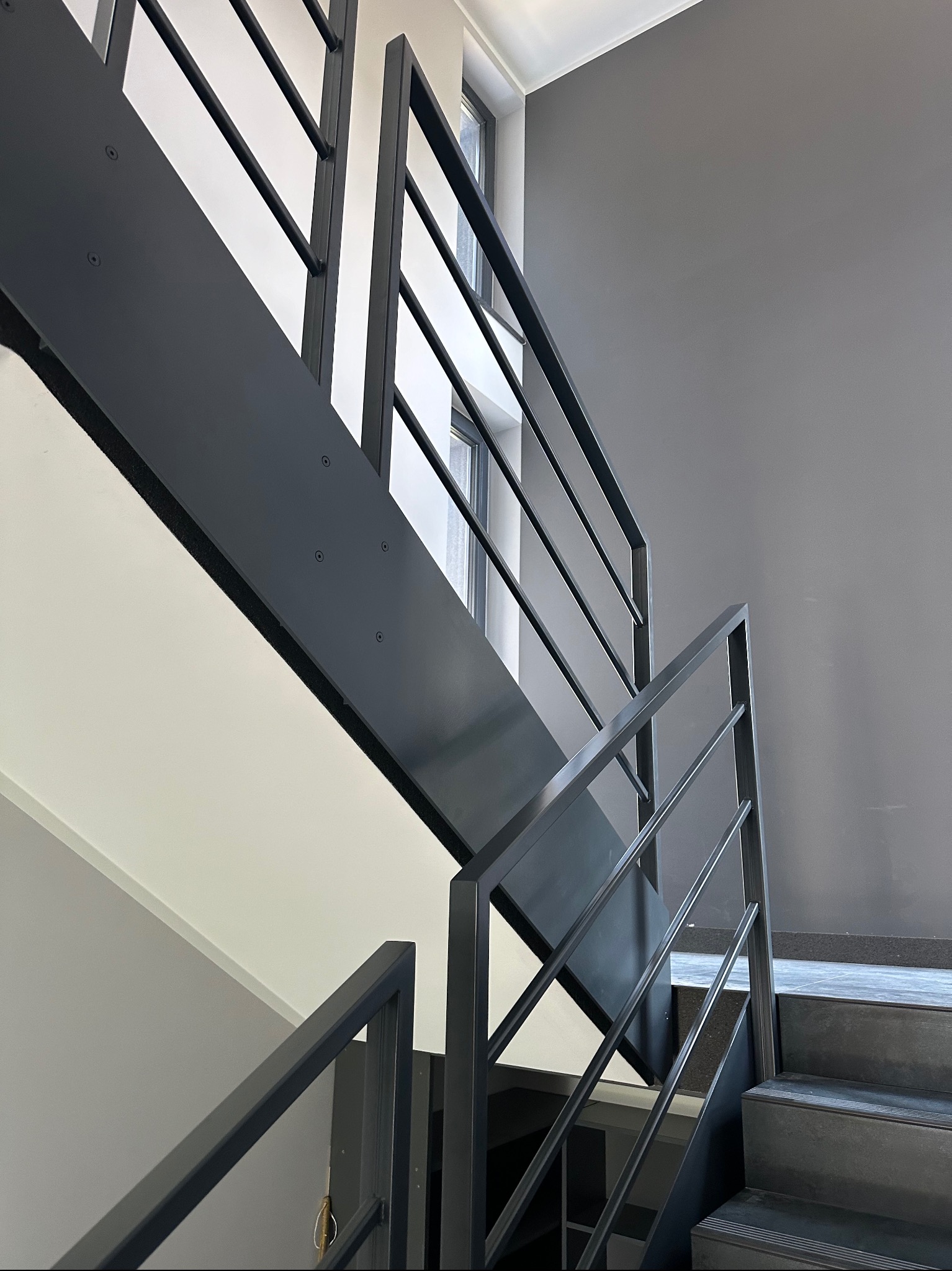 Nowoczesne schody z metalową poręczą w kolorze grafitowym, widok z dołu, minimalistyczne wnętrze z szarą ścianą i oknem w tle.