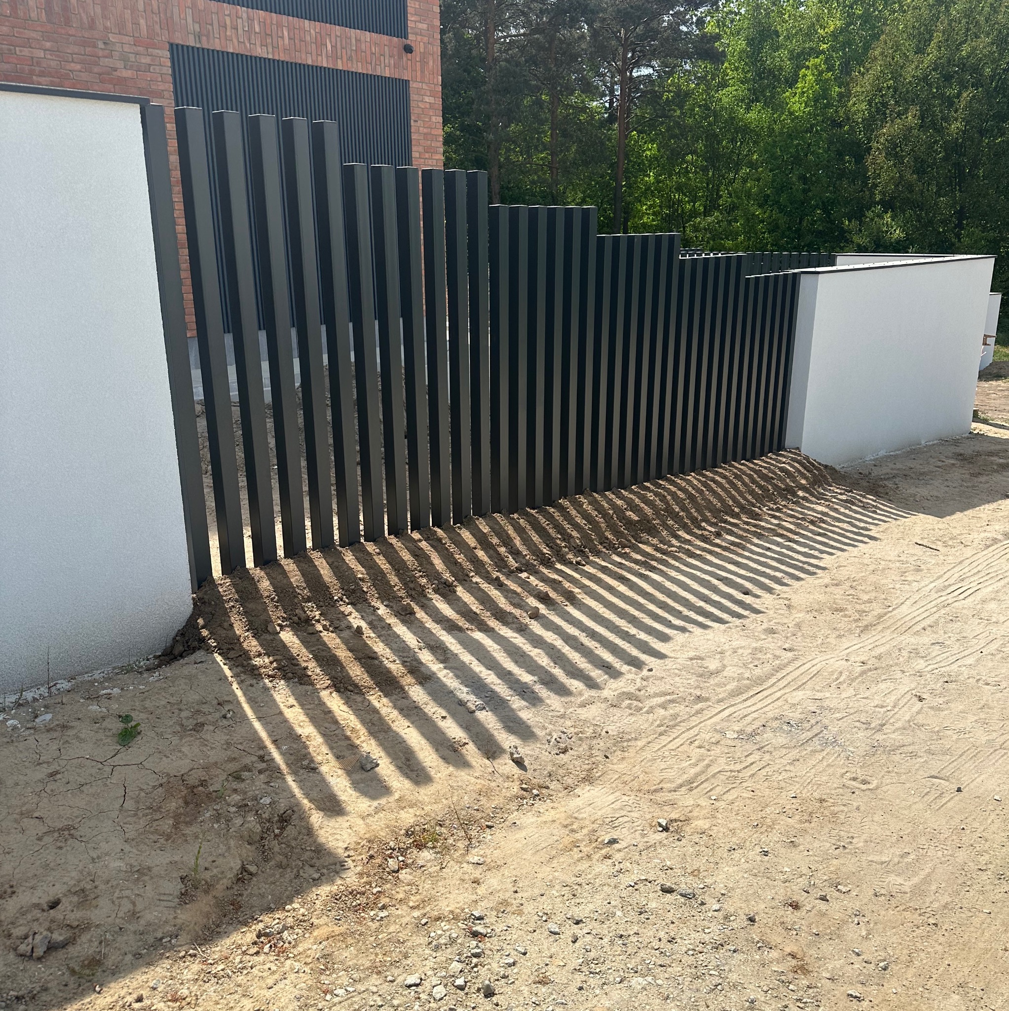 Nowoczesne ogrodzenie panelowe z pionowych, grafitowych profili, osadzone między białymi, murowanymi słupkami. Cienie rzucane przez panele na piaszczystym podłożu.
