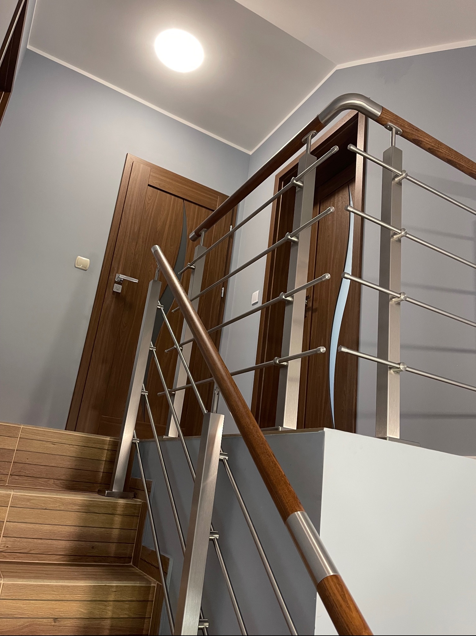 Wnętrze z drewnianymi schodami wyłożonymi płytkami imitującymi drewno i nowoczesną balustradą ze stali nierdzewnej z drewnianym pochwytem, widok z dołu na drzwi w tle.