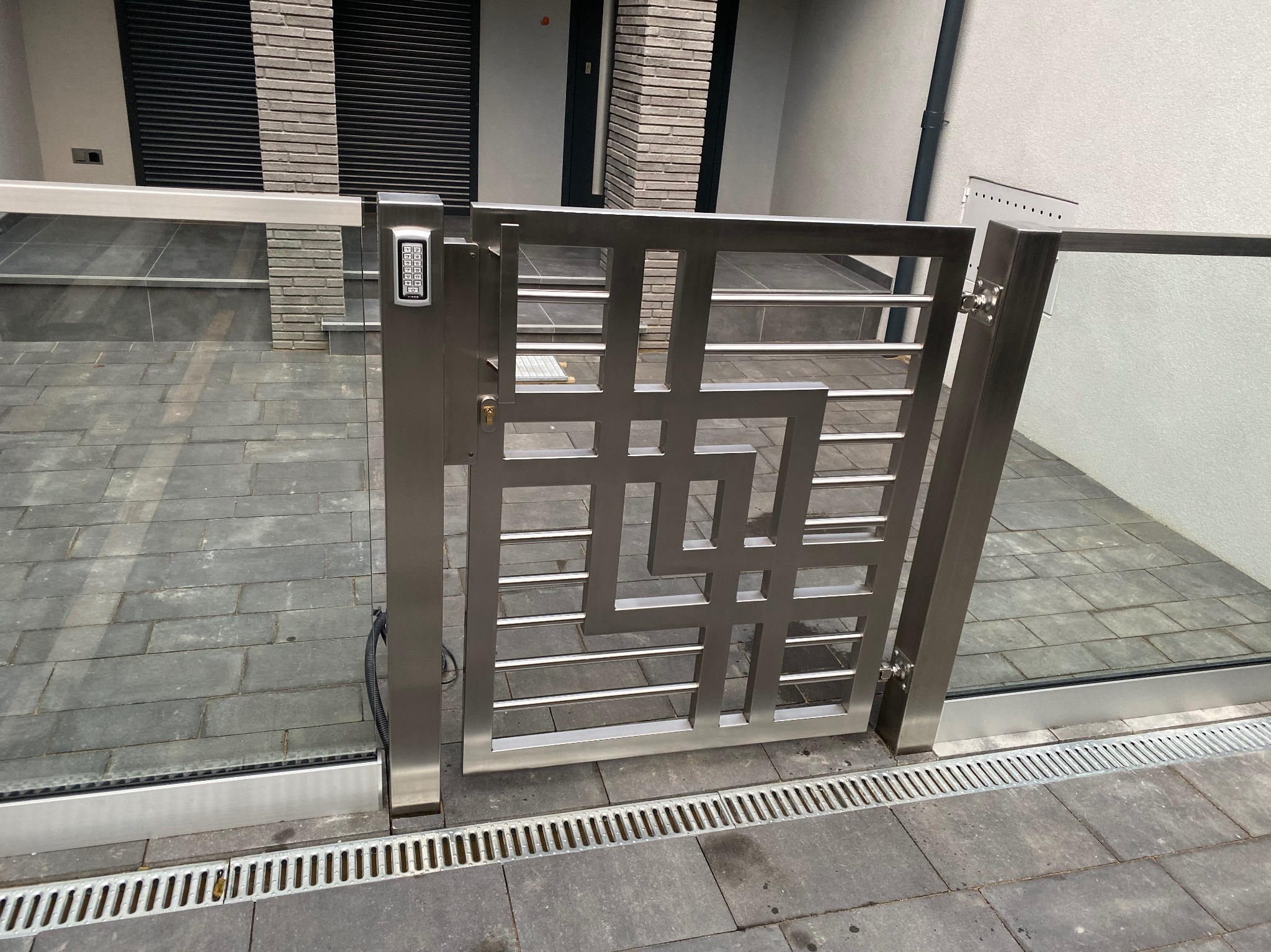 Nowoczesna brama ze stali nierdzewnej z panelem dostępu numerycznego, wkomponowana w szklane ogrodzenie na tle szarej kostki brukowej i jasnej elewacji budynku.