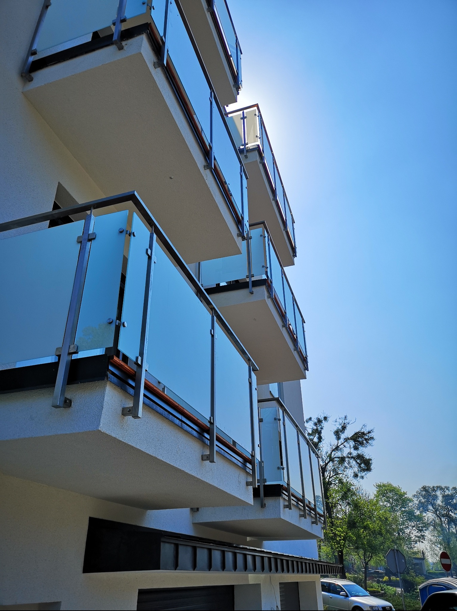 Nowoczesny budynek z balkonami zabezpieczonymi balustradami szklanymi na metalowych wspornikach, widok z dołu w słoneczny dzień.