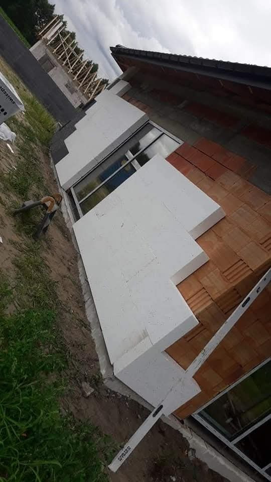 Fragment ściany budynku w trakcie prac: białe płyty izolacyjne przyklejone do muru z czerwonej cegły, widoczny poziomica i okno, w tle konstrukcja drewniana