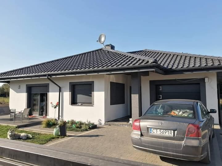 Jednopiętrowy dom z ciemną dachówką, szarą stolarką okienną i elewacją w kolorze złamanej bieli, na podjeździe stoi szary samochód osobowy, a na trawniku leży pies rasy husky.